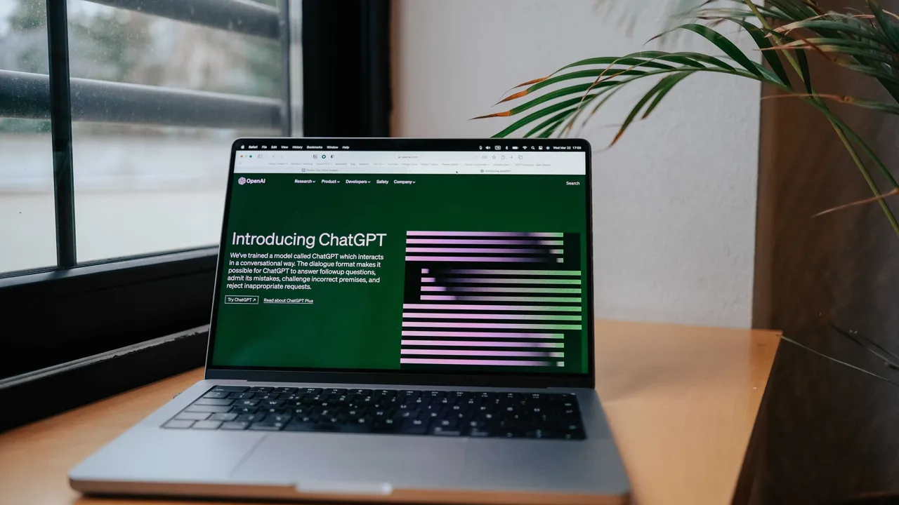 A laptop open on a wooden desk beside a window and a houseplant, showing OpenAI's 'Introducing ChatGPT' announcement page on screen